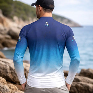 Um pescador visto de costas e usando a Camisa de Pesca Extreme na cor Blue Sea e usando também um boné preto atrás dele tem uma costeira de mar desfocada