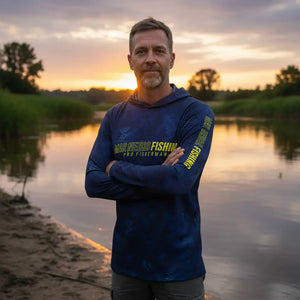 Pescador visto de frente com os braços cruzados usando a Camisa de Pesca Mar Negro Sublimada Capuz Blue, ao fundo um rio desfocado