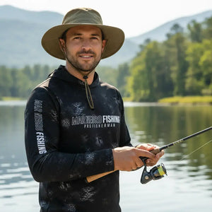 Um pescador com um chapéu visto de frente as margens de um rio segurando uma vara de pesca nas mãos, usando a Camisa de Pesca Mar Negro Sublimada Capuz Black