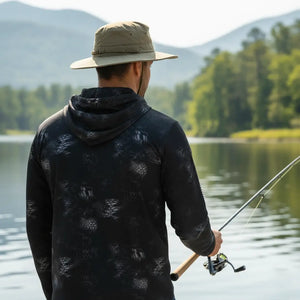 Pescador com um chapéu visto de costas segurando uma vara de pesca e usando a Camisa de Pesca Mar Negro Sublimada Capuz Black, ao fundo tem um rio desfocado