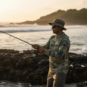 Um pescador visto de lado, segurando uma vara de pesca e usando um chapéu, um óculos e a Camisa de Pesca Mar Negro Sublimada Capuz Caqui, ao fundo o mar em desfoque