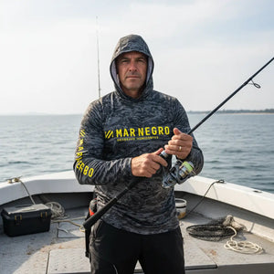 Pescador com capuz visto de frente em cima de uma barco segurando uma vara de pesca, usando a Camisa de Pesca Mar Negro Sublimada Capuz Chumbo