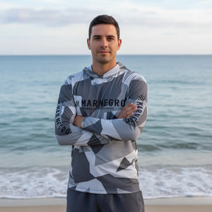 Pescador visto de frente com os braços cruzados usando a Camisa de Pesca Mar Negro Sublimada Capuz Geométrica, ao fundo o mar desfocado