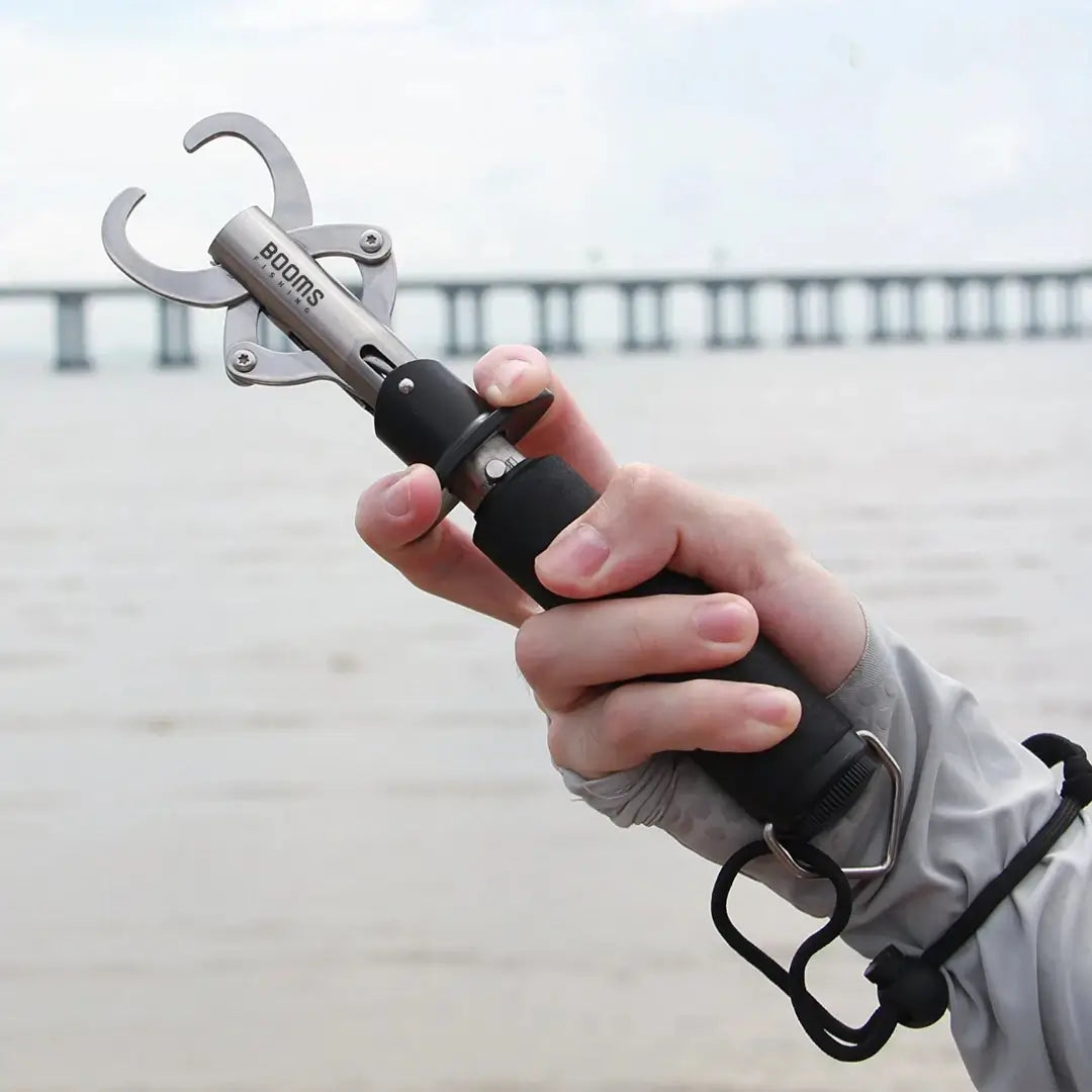 Uma mão segurando o Alicate de Contenção Booms Gripper, puxando seu gatilho e com as pinças abertas, atrás tem um mar em desfoque