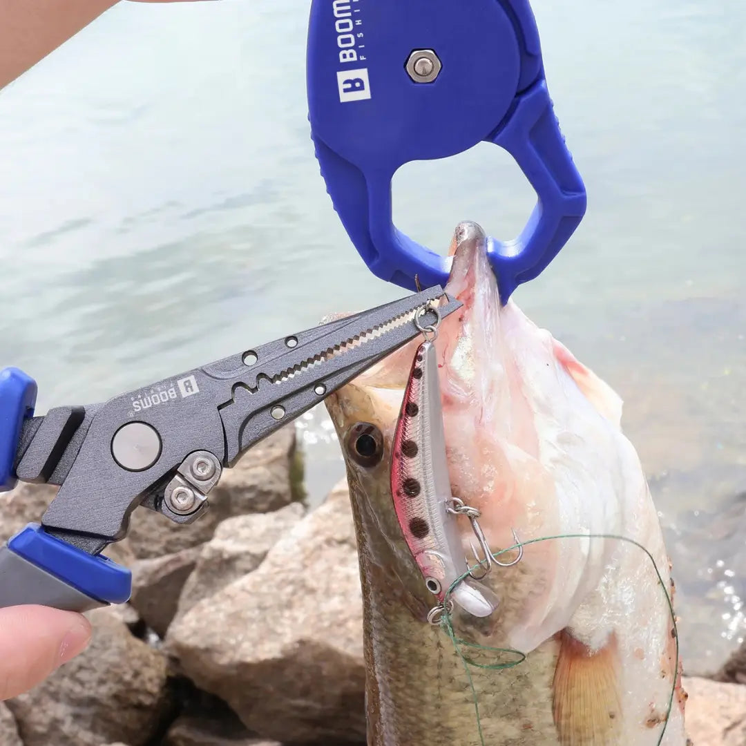 Um peixe segurado por um alicate de contenção, enquanto o Alicate de Pesca Multifunções Booms remove uma isca artificial da boca do peixe
