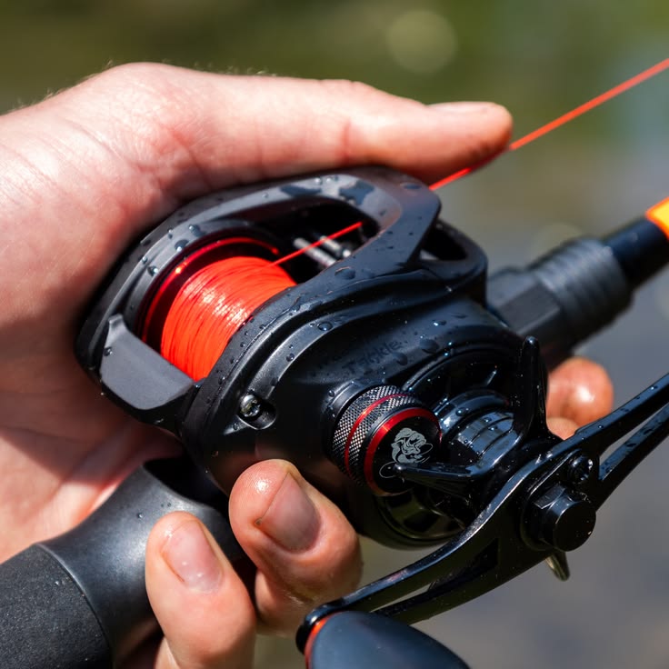 Close em um pescador segurando uma carretilha de pesca na mão, ela tem a cor preta e está usando uma linha multifilamento na cor laranja