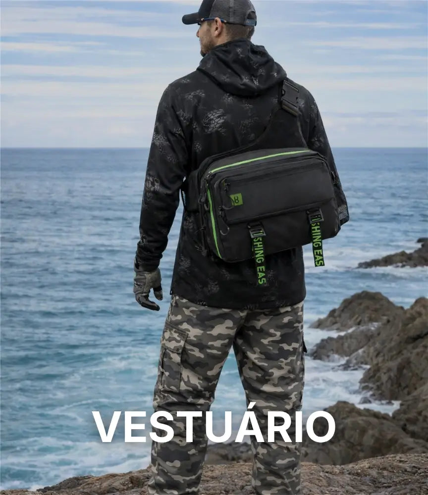 Um pescador usando uma camisa, calça, luvas e chapéu de pesca, ele está na costeira de uma praia e está segurando uma vara de pesca na mão