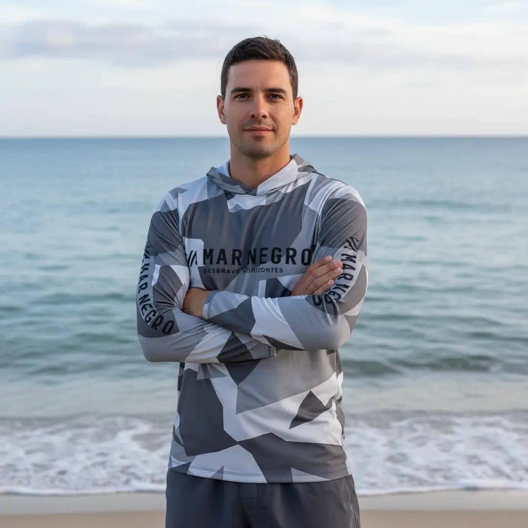 Pescador visto de frente com os braços cruzados usando a Camisa de Pesca Mar Negro Sublimada Capuz Geométrica, ao fundo o mar desfocado