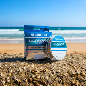 Caixa e o carretel da Linha Monofilamento Shimano Ultegra Invisitec em cima de uma pedra à beira de uma praia ao fundo